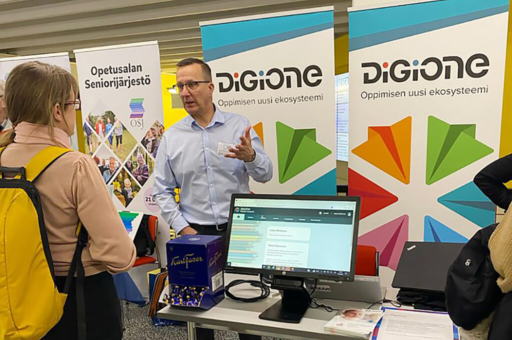  Mies esittelee yleisölle DigiOnea messuständillä, jossa on esillä palvelun demo tietokoneen näytöllä sekä messuesitteitä ja rollup-julisteita.